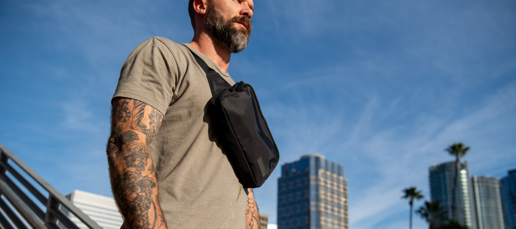 A man with tattooed arms wears a black Faraday sling by SLNT across his chest against a city skyline. The sling bag features a built-in Faraday cage for signal blocking.